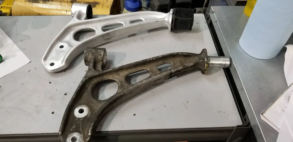 Worn suspension control arm beside a new one on the workbench at Alpine Autowerks